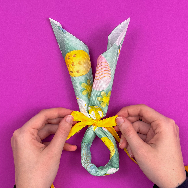 Ein Bild das zeigt, wie mit dem Geschenkband eine Schleife um die Serviette gebunden wird.