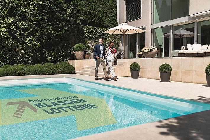 Ein Paar schlendert im Garten einer Luxusvilla entlang eines großen Pools, im Pool ist das Logo der Klassenlotterie zu sehen.