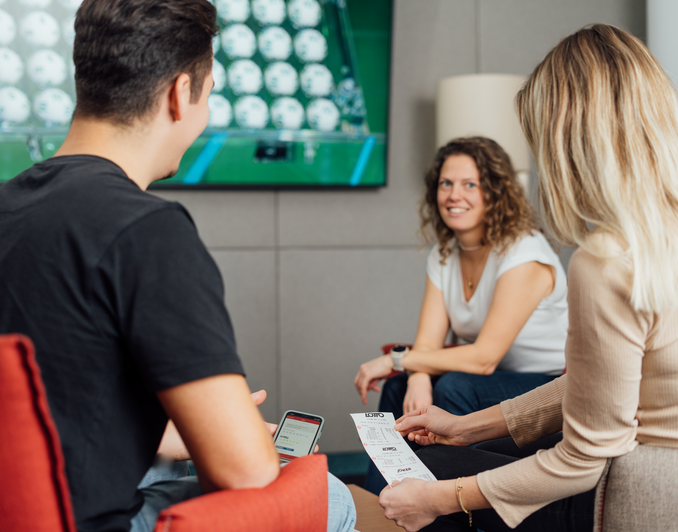 Drei junge Menschen sehen sich die Lotto-Ziehung im Fernsehen an und vergleichen ihre gespielten Lotto-Zahlen