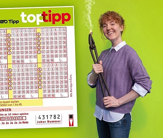 Eine junge Frau hält eine glühend heiße Euromünze mit einer Eisenzange. Daneben ein Top-Tipp-Schein, ein Zahlen-Spiel, bei dem man auf die Lotto-Zahlen tippt.